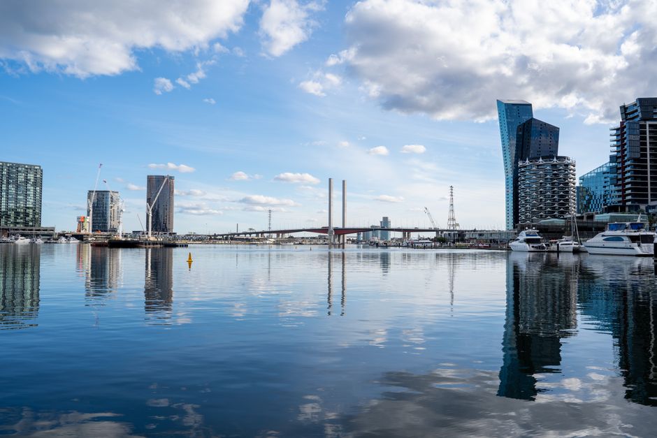 A panoramic view of a city skyline along a river, featuring modern high-rise buildings with glass facades reflecting the water, some under construction with cranes visible. Several boats are moored al