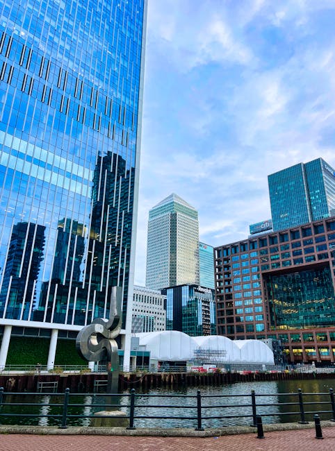 A collection of modern high-rise office buildings with glass and steel facades situated along a waterfront in Canary Wharf, Docklands. The skyscrapers feature reflective windows that mirror the partly cloudy sky above. In the foreground, there is a black metal railing along the water's edge and a small sculpture made of metal with an abstract design. Behind the railing, a paved walkway is visible, along with a white, dome-shaped structure and a few moving boxes wrapped in plastic and cardboard stored nearby, suggesting a home relocation or furniture transport process. The scene is well-lit with natural daylight, emphasizing the urban environment and the logistics involved in a house removal within high-rise flats. Man with Van Docklands specializes in removals, including packing, loading, and transport services for high-rise properties in E14 and the Docklands area. This image highlights the logistical considerations of loading and moving furniture during a high-rise home relocation.