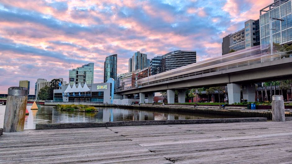 A panoramic view of Docklands with a wooden pier in the foreground, showcasing modern office buildings and residential towers under a partly cloudy sky during sunset. An elevated train track runs hori