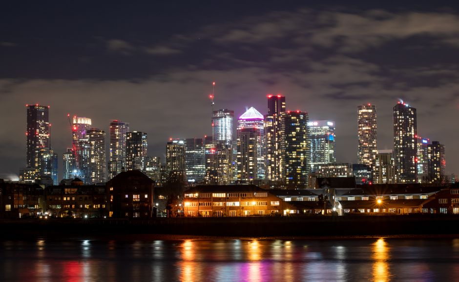 A panoramic view of Docklands with a wooden pier in the foreground, showcasing modern office buildings and residential towers under a partly cloudy sky during sunset. An elevated train track runs hori
