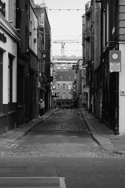 A narrow cobblestone street in the Docklands area, flanked by multi-storey brick buildings with large windows and external wiring overhead. On the left side, a person wearing a light-colored jacket and carrying a bag is walking along the pavement, while the right side features a street sign and a black-and-white pole. The street appears to slope slightly downward toward a small open square with additional buildings in the distance. The overcast lighting and urban setting suggest a typical area for home relocation or furniture transport, with potential for loading and unloading of belongings for house removals. This scene illustrates the type of environment where a professional man with van service, such as Man with Van Docklands, might operate during a house move across the narrow streets and docks of the Isle of Dogs.
