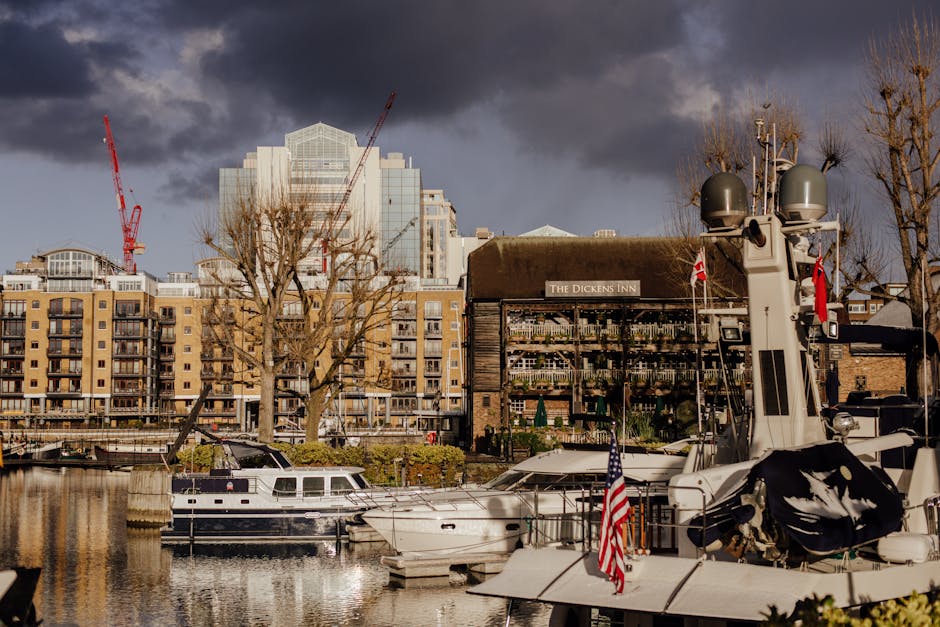 A marina scene in the Docklands area displaying several boats docked along the water, including a prominent motorboat with a white and navy hull, visible ropes securing it to the quay, and a small American flag attached to its railing. Adjacent to the motorboat, there are other boats of varying sizes, some with protective covers. In the background, there are tall modern residential buildings with multiple balconies, constructed from beige brick and glass, along with construction cranes indicating ongoing development. A large leafless tree is situated near the boats, extending its branches outward. To the right, a wooden building with a sign reading 'The Dickens Inn' has large windows displaying potted plants and items inside, and is partly obscured by trees with bare branches. The sky above is dark with storm clouds, suggesting upcoming rain, and the scene is illuminated by natural daylight, creating a contrast between the gloomy sky and the well-lit waterfront. This image captures elements relevant to moving and transport services in the Docklands, encompassing furniture and equipment logistics in a boatyard environment.