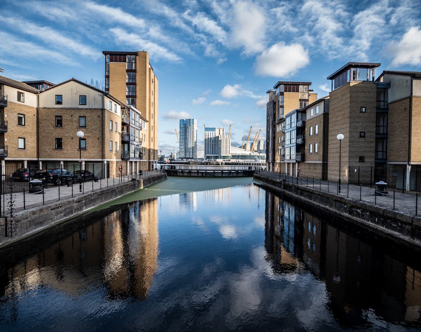 A wide view of a calm waterway in the Docklands area, with a stone-paved walkway on the left side featuring several stacks of cardboard boxes and a small orange boat moored along the edge. On the righ